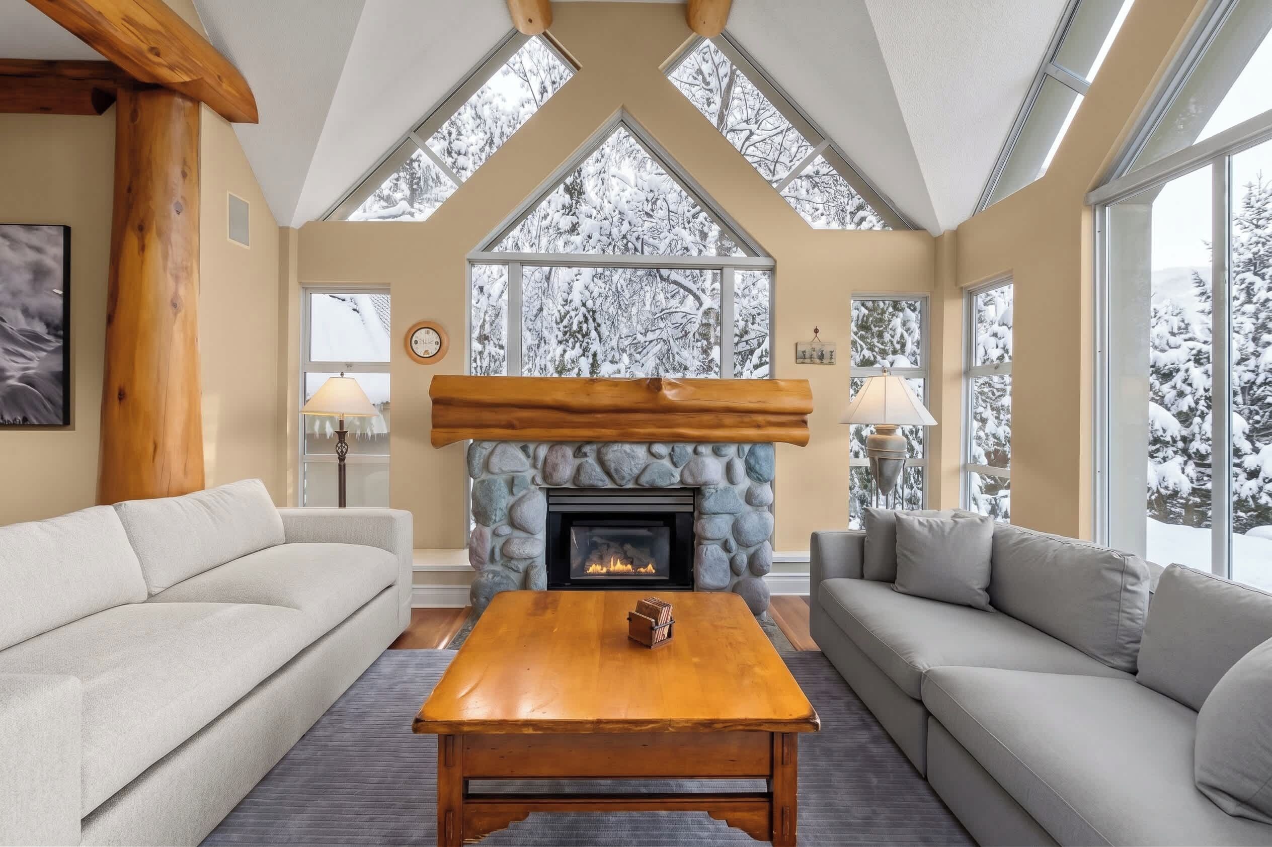 A cozy living room with a stone fireplace, wooden beams, and large windows with snowy views.