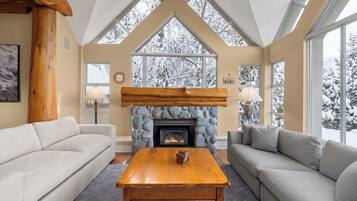 A cozy living room with a stone fireplace, wooden beams, and large windows with snowy views.