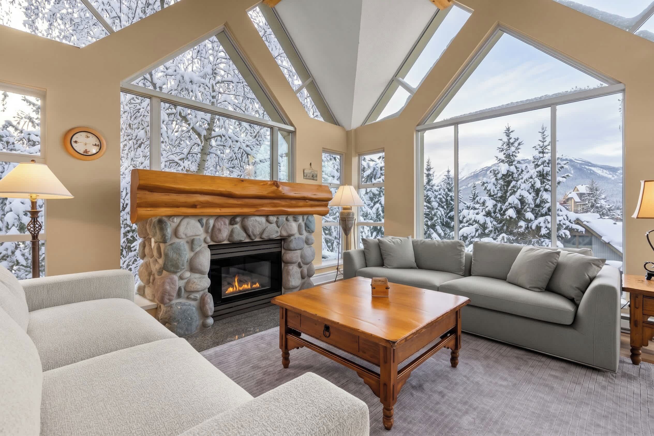A cozy living room with a stone fireplace, large windows, and a view of snow-covered trees.