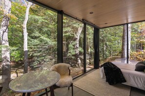 Una habitación con un gran ventanal, una butaca blanca de pelo, una mesa de vidrio y una cama con una colcha negra.
