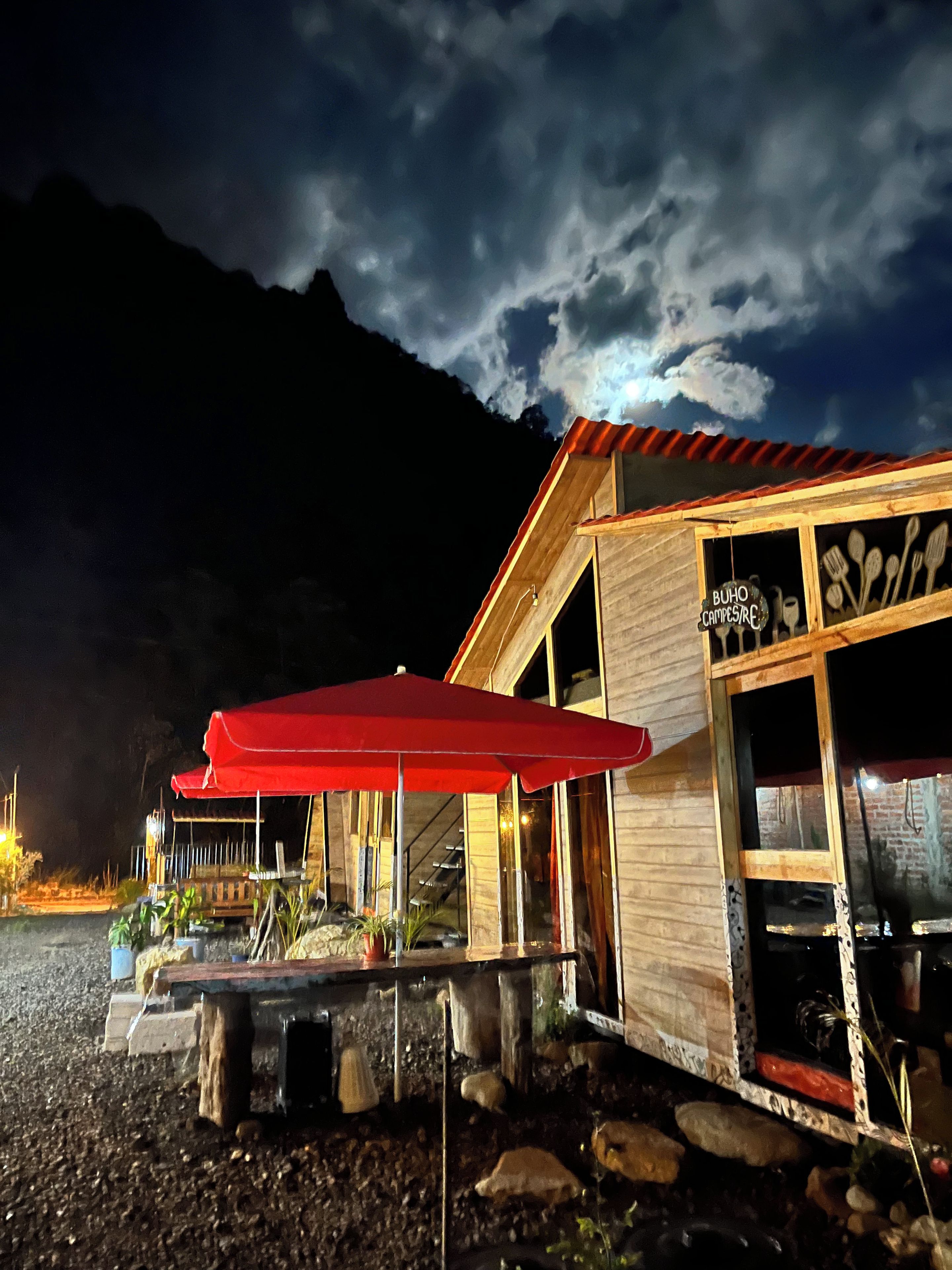 Un bâtiment en bois surmonté d’un parasol rouge, une enseigne indiquant « BUHO CAMPS/RESTAURANT » et, en arrière-plan, une vue sur des montagnes.