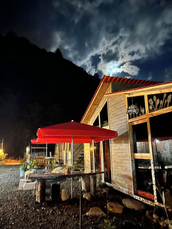 A wooden building with a red umbrella, a sign reading 'BUHO CAMPS/RESTAURANT', and a view of mountains in the background.