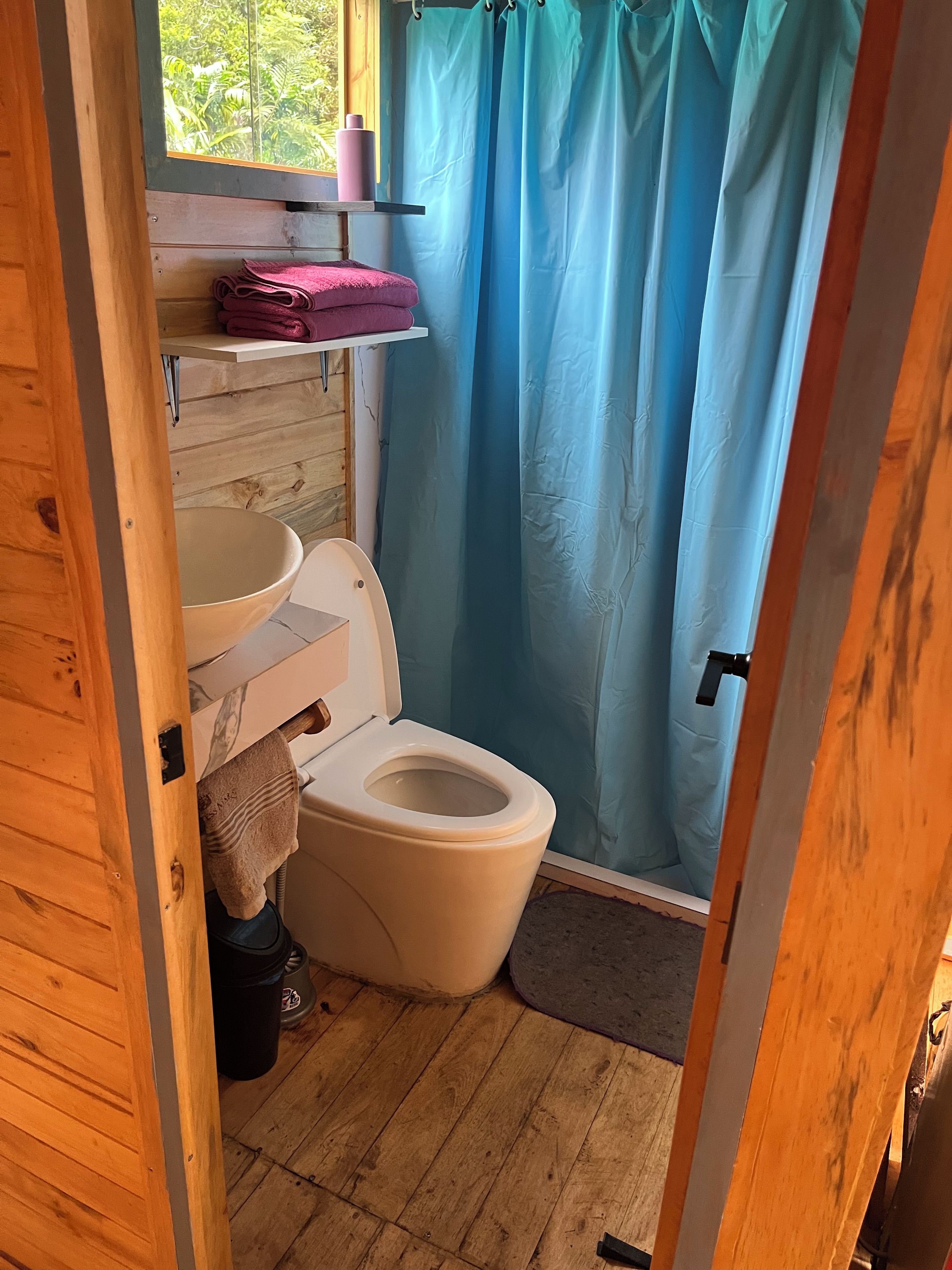 Une petite salle de bain avec un plancher en bois, des toilettes blanches, une étagère en bois sur laquelle est posée une serviette rose, et un rideau de douche bleu.