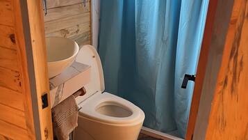 Une petite salle de bain avec un plancher en bois, des toilettes blanches, une étagère en bois sur laquelle est posée une serviette rose, et un rideau de douche bleu.