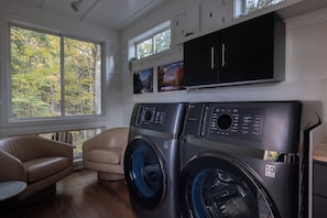 A laundry room with two front-loading washing machines, a beige sofa, and a large window with a view of trees. - Wildflower | Mirror Cabin + Spa at Uncommon Accommodations - Smuggler's Notch (Cambridge)