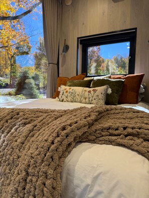 A bedroom with a large window, a bed with a brown knitted blanket, and multiple pillows. - Wildflower | Mirror Cabin + Spa at Uncommon Accommodations - Smuggler's Notch (Cambridge)