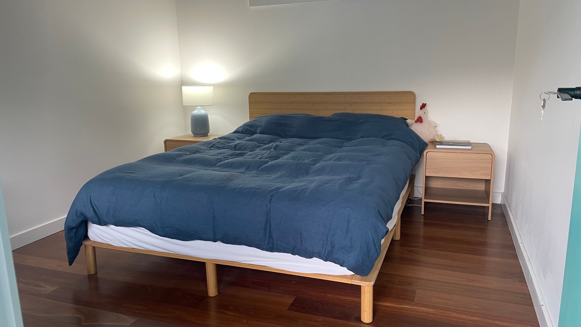 Un lit bien fait, avec une couette bleue et des draps blancs, une lampe de chevet et une petite table de nuit en bois sur laquelle se trouve une autre lampe.