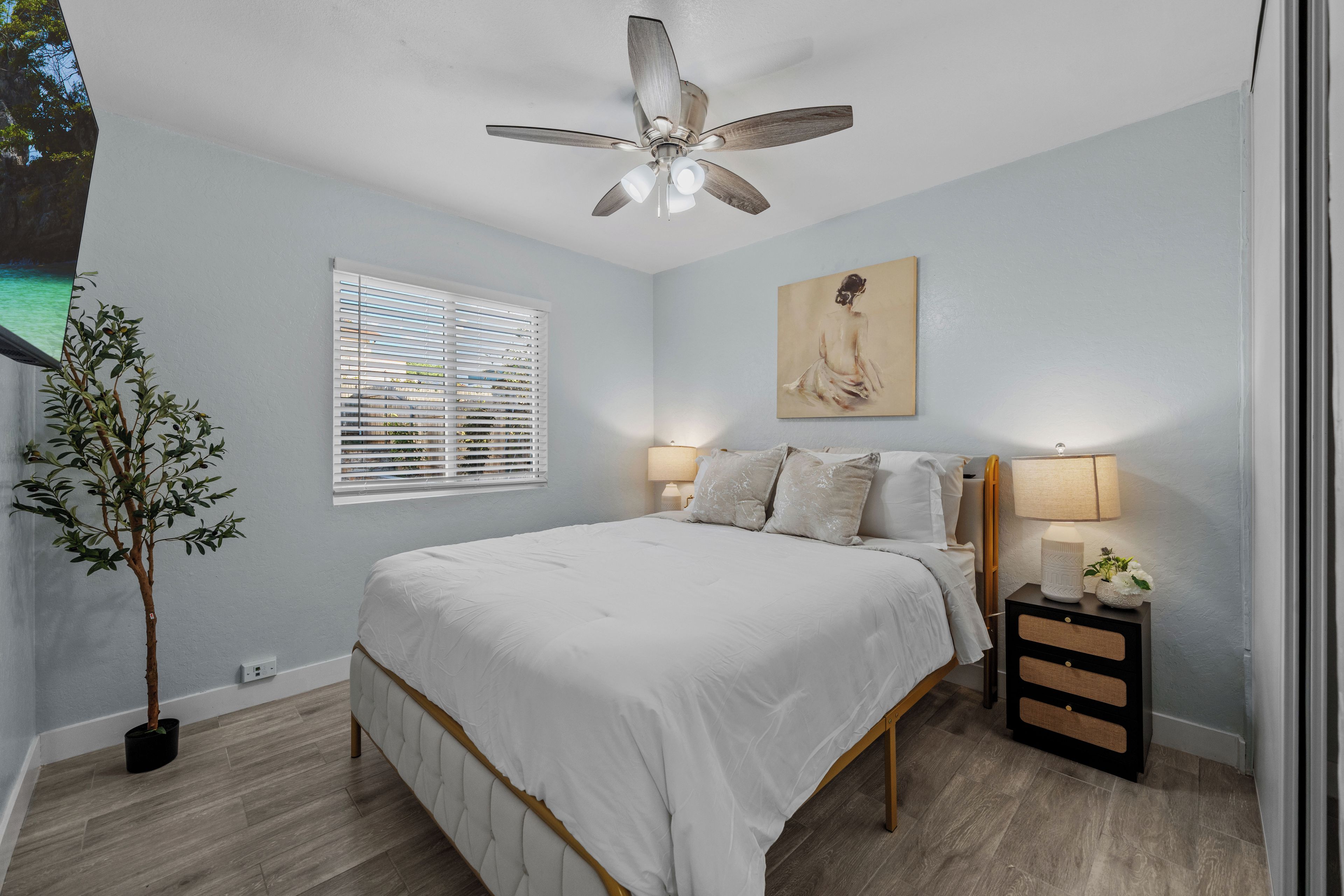 Une chambre à coucher comprenant un lit, un ventilateur de plafond, des lampes de chevet, une table de chevet, une plante en pot et une fenêtre avec des stores.