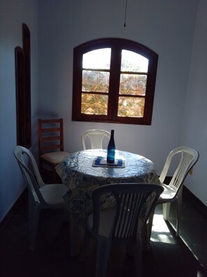 A round table with a patterned tablecloth, surrounded by white plastic chairs, a blue bottle, and a window with a view of trees. - SPECIAL OFFER: House in Itaúna, 3 bedrooms, 2 bathrooms, near the beach, pets welcome. (Saquarema)