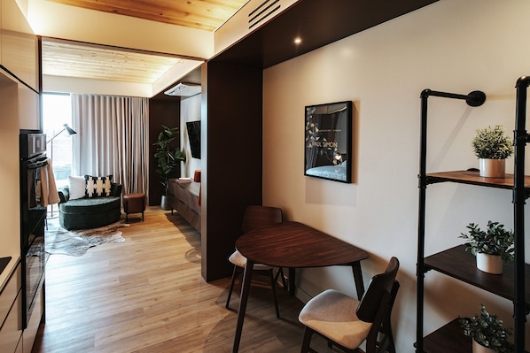 Una sala de estar moderna con suelo de madera, un sofá oscuro, una mesa de comedor con sillas, un televisor y una planta.