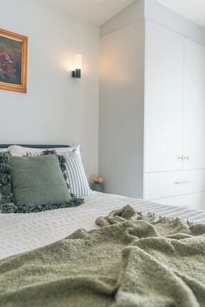 Une chambre à coucher avec un lit, des oreillers, une couverture, une lampe fixée au mur et un tableau.