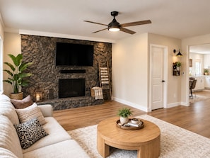 A living room with a stone fireplace, a flat-screen TV, a ceiling fan, and a wooden coffee table.