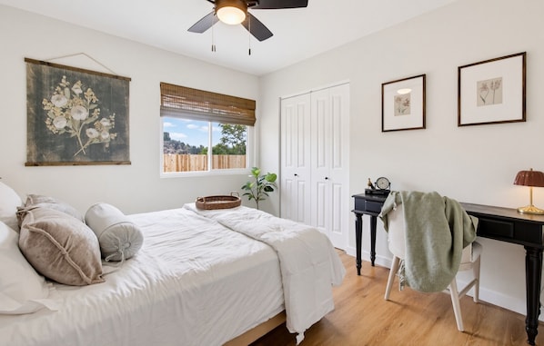 A bedroom with a bed, a desk, a chair, a ceiling fan, and a window with blinds.
