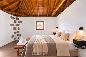 A bedroom with a wooden ceiling, a stone wall, a bed with a quilted cover, bedside tables with lamps, and a window with a view. - TenoHomes Villa SAYAN, Private Pool & Teide Views (Icod de los Vinos)