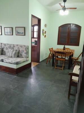 A living room with a sofa, a dining table with chairs, a ceiling fan, and a window with a wooden frame. - House in Itauna with 3 bedrooms, 1 suite, balcony, near the beach, parking for 3 cars, green area. (Saquarema)