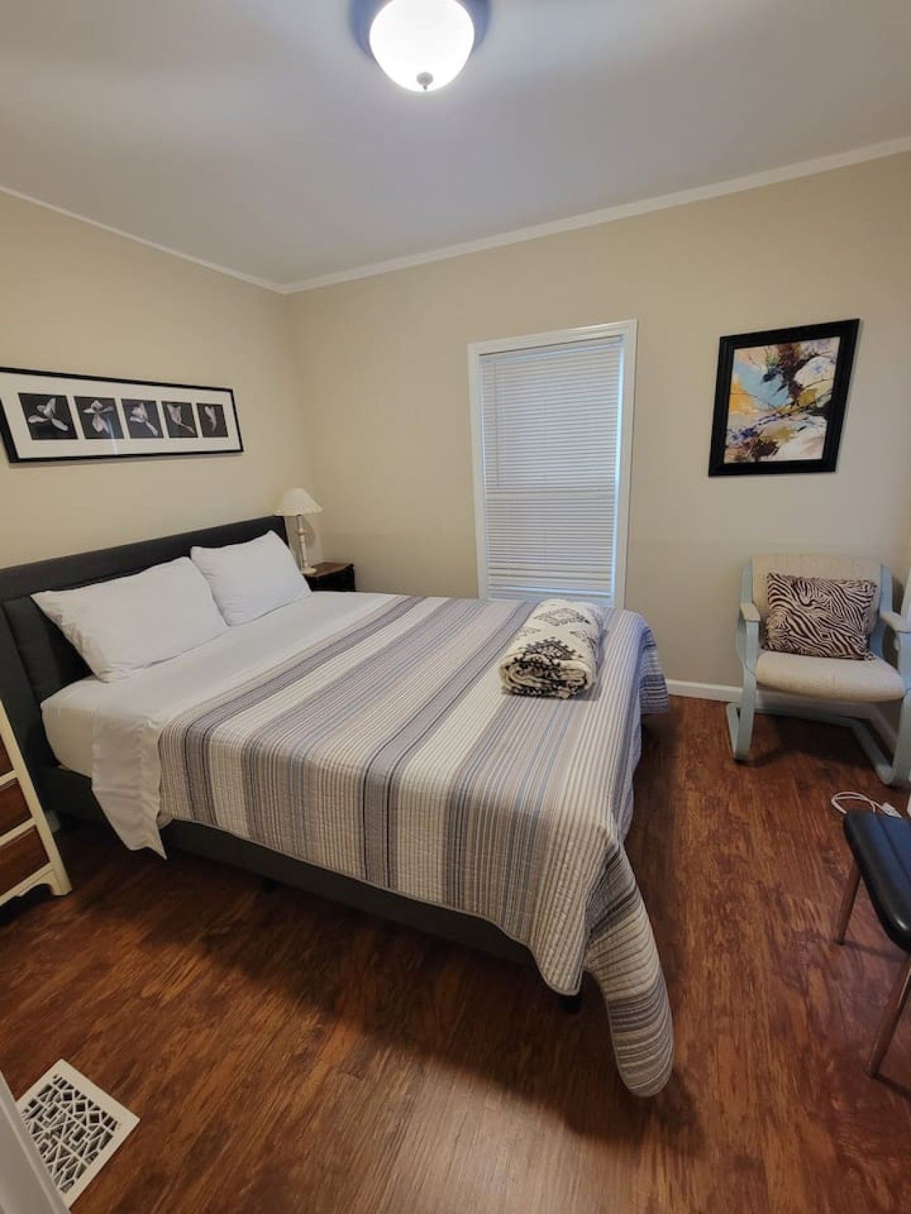 A bedroom with a bed, a chair, a window with blinds, a wall with framed pictures, and a ceiling light.