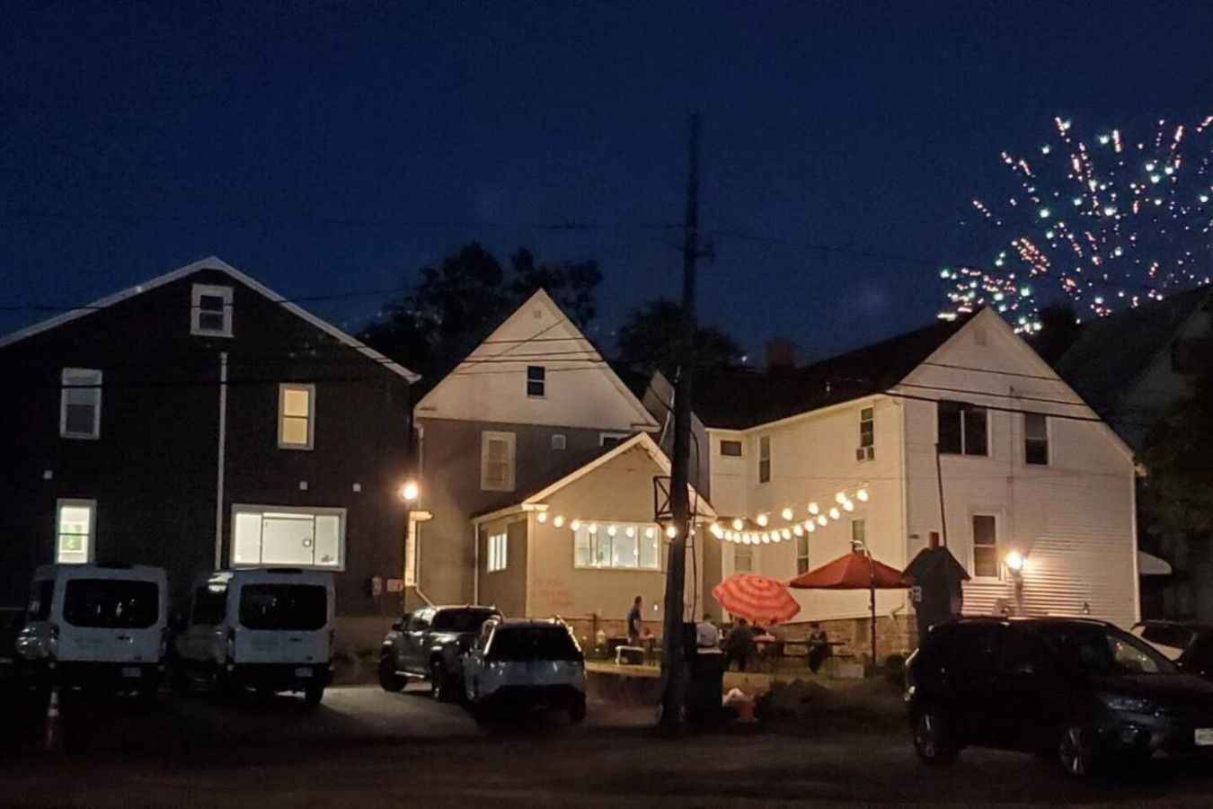 A street view at dusk with houses, parked cars, and a visible firework display in the sky.