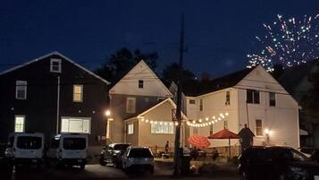A street view at dusk with houses, parked cars, and a visible firework display in the sky.