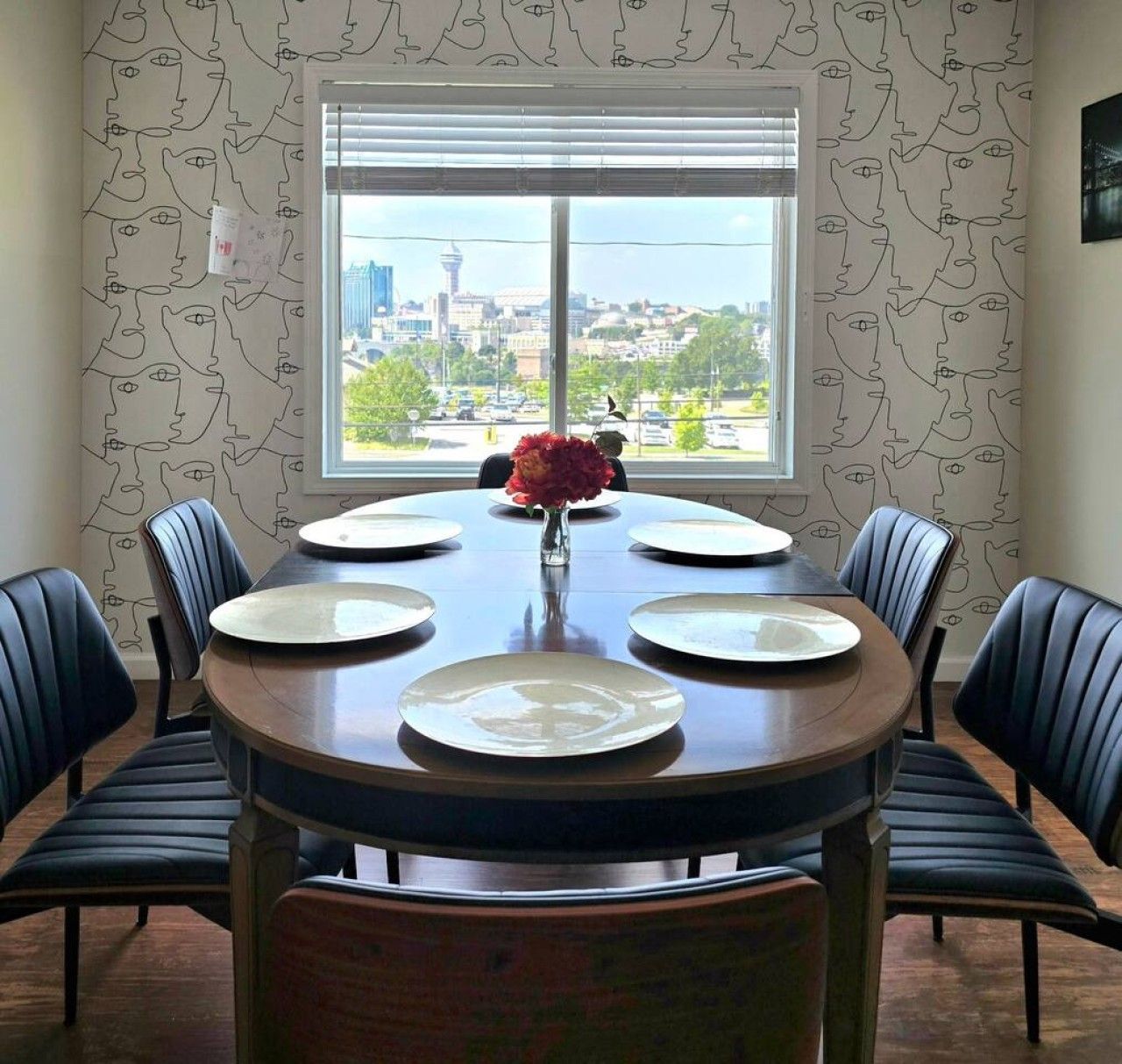 A dining room with a round table set for six, flanked by six chairs, and a window with a cityscape view.