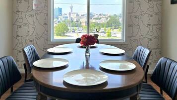 A dining room with a round table set for six, flanked by six chairs, and a window with a cityscape view.