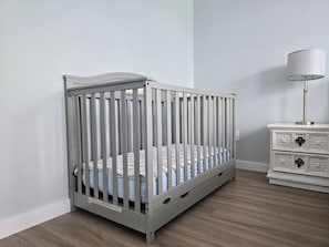A white crib with vertical slats, a white nightstand with drawers, and a floor lamp in a room with light-colored walls and wooden flooring. - “Life is Good” | Accessible Stay Near the Falls (Niagara Falls)