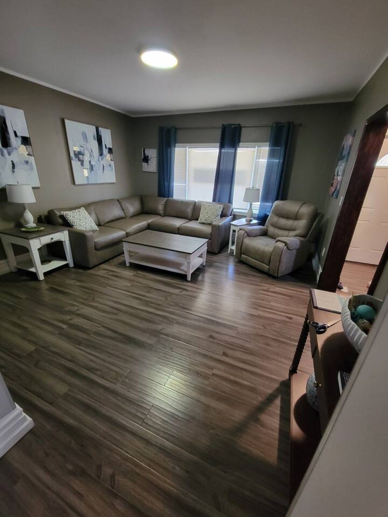 A living room with a sectional sofa, a coffee table, and a chair. There are two lamps on side tables, and artwork on the walls.