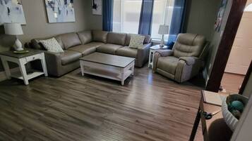 A living room with a sectional sofa, a coffee table, and a chair. There are two lamps on side tables, and artwork on the walls.