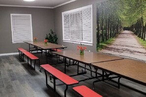 Ein Konferenzraum mit langem Tisch und Bänken, großen Fenstern mit Jalousien und einem Wandbild von einem Baumweg.