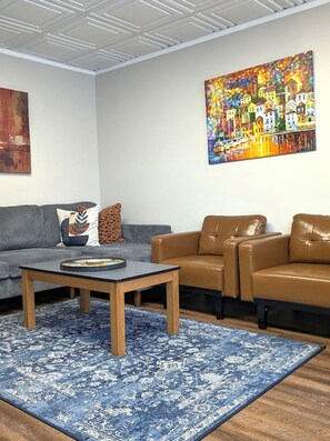 A living room with a sofa, a coffee table, and a brown leather chair. There is a colorful painting on the wall and a blue patterned rug on the floor. - Gray Goose - Walk to Falls | Spacious 4BR Home (Niagara Falls)
