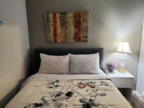 A hotel room with a bed, a nightstand, a lamp, and a painting on the wall. - Gray Goose - Walk to Falls | Spacious 4BR Home (Niagara Falls)