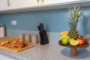 A kitchen counter with a fruit and pastry display, a knife block, and a blue tiled backsplash. - Greater Manchester | Whole home | 5-Bedroom | Sleeps 8 | (Tyldesley)