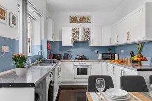 A modern kitchen with white cabinets, a granite countertop, a built-in oven, and a dining area with a table set for two. - Greater Manchester | Whole home | 5-Bedroom | Sleeps 8 | (Tyldesley)