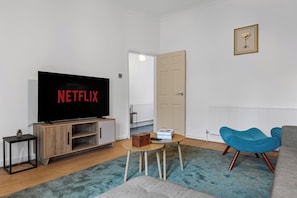 A living room with a flat-screen TV displaying 'NETFLIX,' a wooden entertainment unit, a small table with a book and a remote, a blue chair, and a grey sofa. - Greater Manchester | Whole home | 5-Bedroom | Sleeps 8 | (Tyldesley)