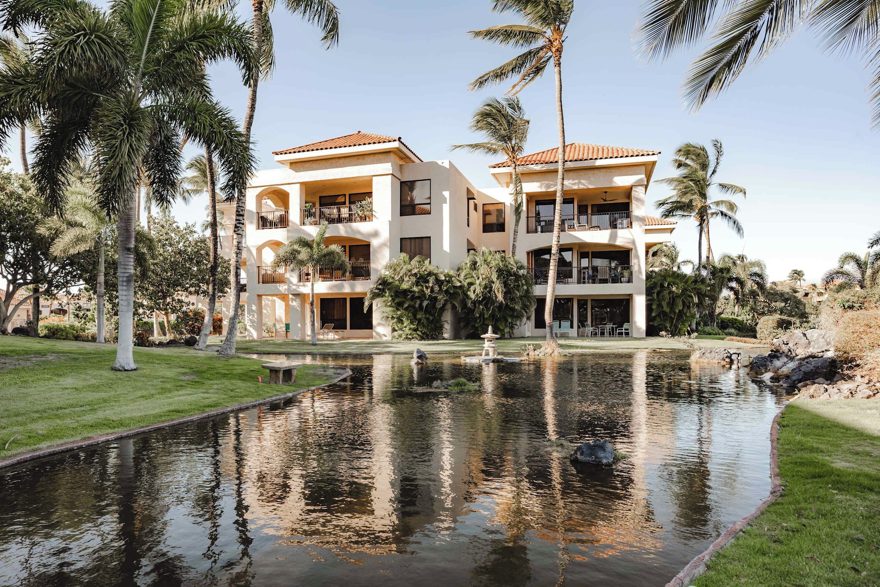 A two-story building with balconies, surrounded by palm trees and a pond.