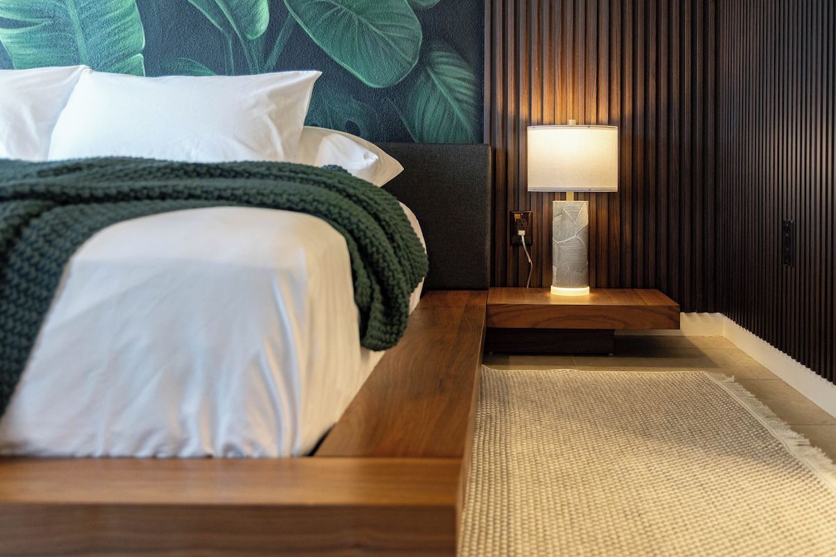 A neatly made bed with white linens and a green throw blanket, a bedside lamp, and a textured rug on a wooden floor.