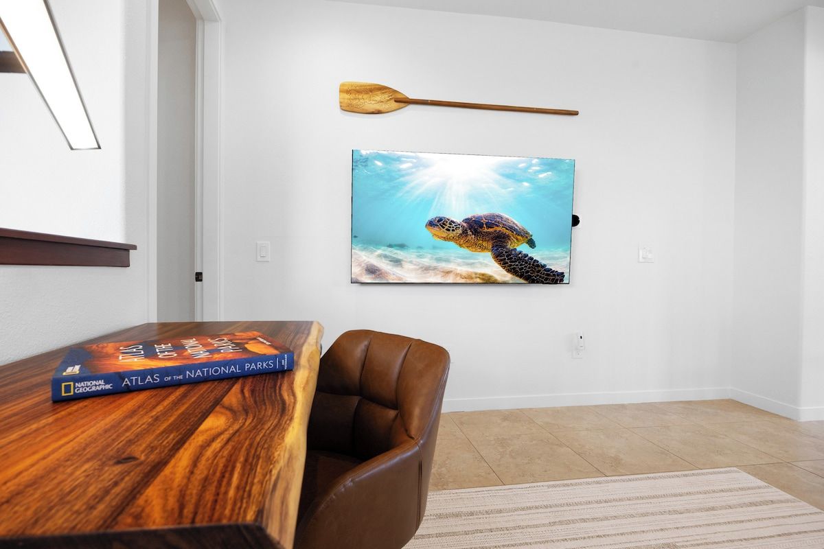 A room with a wooden table, a brown leather chair, a framed picture of a sea turtle, and a book titled 'Atlas of the National Parks'.