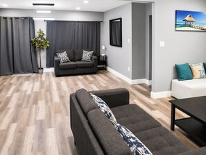 Ein modernes Wohnzimmer mit einem grauen Sofa, einem dunklen Sessel und einem weißen Sofa. Es gibt eine Pflanze, ein gerahmtes Bild an der Wand und Holzboden.