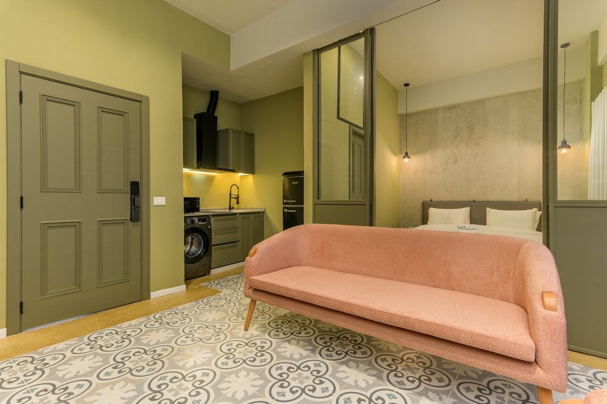 Habitación de hotel compacta con sofá, lavadora, cama y un espejo grande.