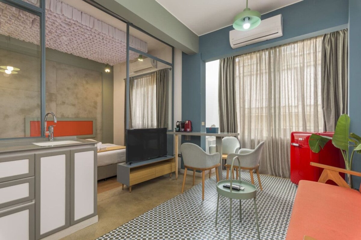 Habitación de hotel compacta con kitchenette, una cama, televisor, zona de comedor con sillas y un gran ventanal con cortinas.