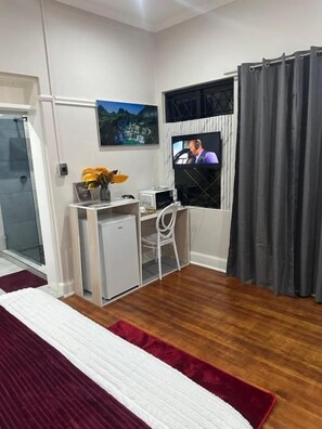 A compact room with a bed, a desk, a chair, a small fridge, and a television. - Comrades villa guest house  (Pietermaritzburg)