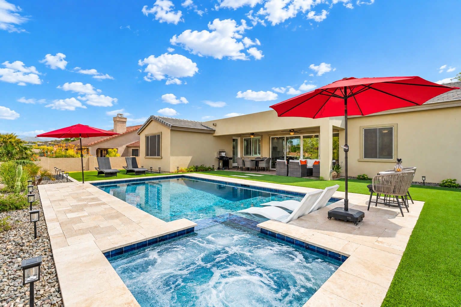 A swimming pool with a hot tub, surrounded by outdoor furniture and umbrellas, set against a clear sky.