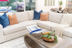 A modern living room with a sectional sofa, a coffee table with a book and decorative items, and a view of the outdoors. - NEW|The Ellington|Pool+Hottub+Game Table|Sleeps 10 (Fountain Hills)