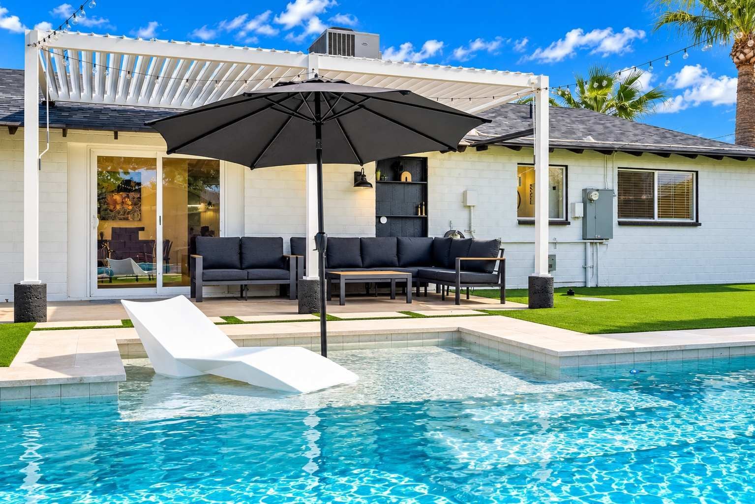 A modern outdoor pool area with a white lounge chair, a black umbrella, and a seating area with sofas and a table.