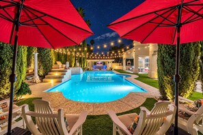 A swimming pool with red umbrellas, surrounded by outdoor seating and a string of lights.