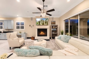 A living room with a fireplace, a ceiling fan, and a large window. - NEW|Lionsgate Luxe Estate | Pool + Spa | Golf Sim (Gilbert)