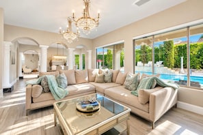 A spacious living room with a large sectional sofa, a glass coffee table, and a pool visible through the large windows.