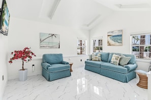 A spacious living room with a high ceiling, a large window with a view of the beach, and a modern blue sofa set. - Turquoise Mermaid Cottage at White Sands | updated 1 br/1ba steps to beach (Indian Rocks Beach)