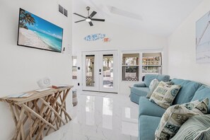 A modern living room with a blue sofa, a wooden coffee table, a ceiling fan, and a beach-themed painting. - Lemon Tree Cottage at White Sands | updated 2br/1ba steps to beach (Indian Rocks Beach)