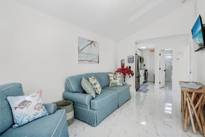 Ein Wohnzimmer mit einem blauen Sofa, einem kleinen Tisch, einem Fernseher und einer Küchenzeile im Hintergrund.