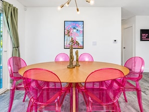 A dining area with a round wooden table surrounded by six pink chairs, a framed artwork above the table, and a wall-mounted sign. - Gorgeous Mountain Sunset Casita with AC Heated Pool Hot Tub Fire pit EV Charger (Palm Springs)